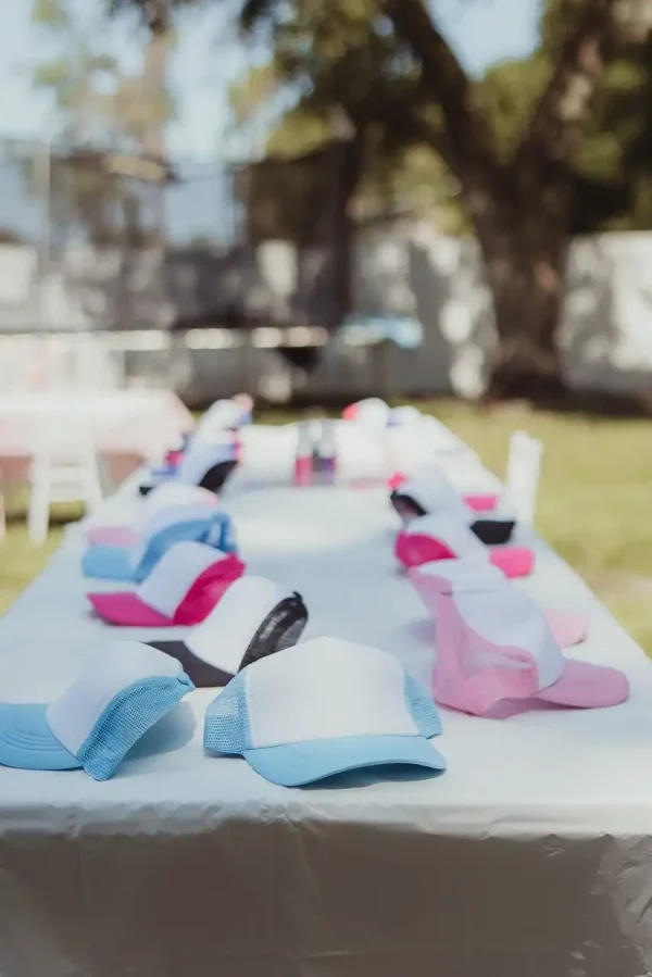 A long table outdoors showcases a vibrant array of trucker hats in blue, pink, white, and black while trees bask in the sunlight in the background. Capture every cherished moment of this colorful scene with our Event Photography Packages.