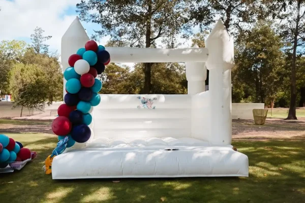 The Event Photography Packages feature a charming white inflatable bounce house adorned with a cluster of multicolored balloons, thoughtfully positioned outdoors on a grassy area with trees in the background, making it ideal for capturing memorable event moments.