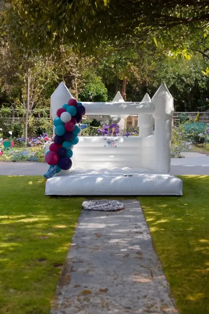 An Event Photography Package features an inflatable white bouncy castle adorned with a delightful cluster of purple, blue, and pink balloons, set on a grassy area with a stone pathway leading up to it. The enchanting backdrop of trees and garden adds the perfect touch for capturing memorable moments.