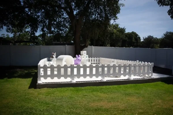 A charming small white picket-fenced area, featuring various animal-shaped decorations and a large tree in the background, set in a grassy yard; perfect for capturing memories with our Event Photography Packages.