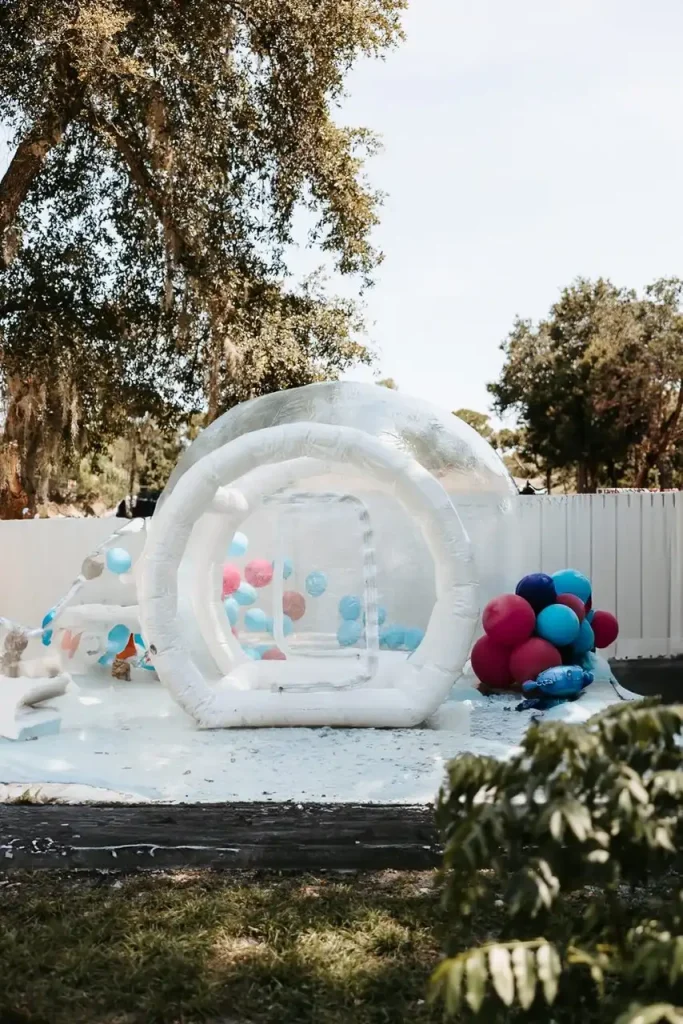 An "Event Photography Package" offers a stunning setting featuring a white inflatable dome filled with colorful balloons, predominantly blue and pink, set up outdoors on a patch of grass. This location is perfect for events or photography sessions, complete with trees and a white fence in the background.