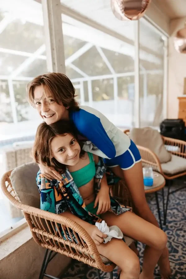 Two children in swimwear indoors, one sitting on a wicker chair and the other leaning over with their arms around the seated child, both smiling. The background includes windows and a pool area, capturing a perfect moment ideal for Event Photography Packages.