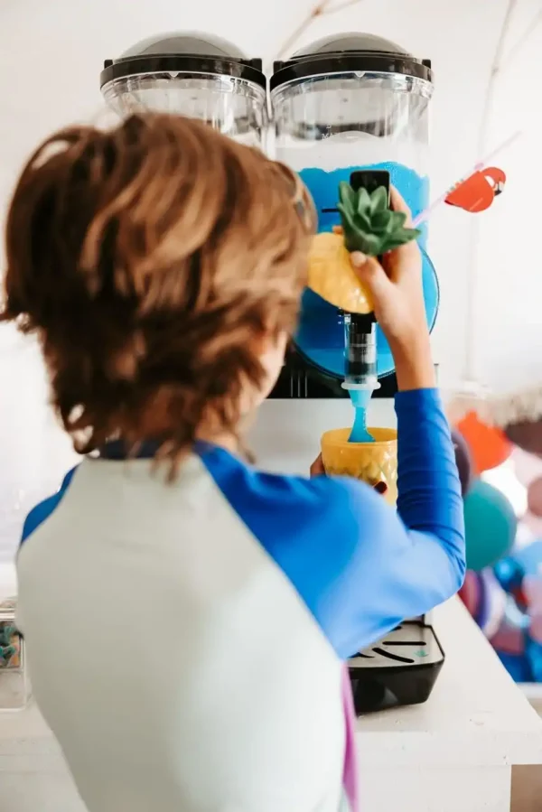 A child with sandy brown hair and a blue shirt fills a pineapple-shaped cup with blue slush from a slush machine, creating the ideal candid moment for our Event Photography Packages.