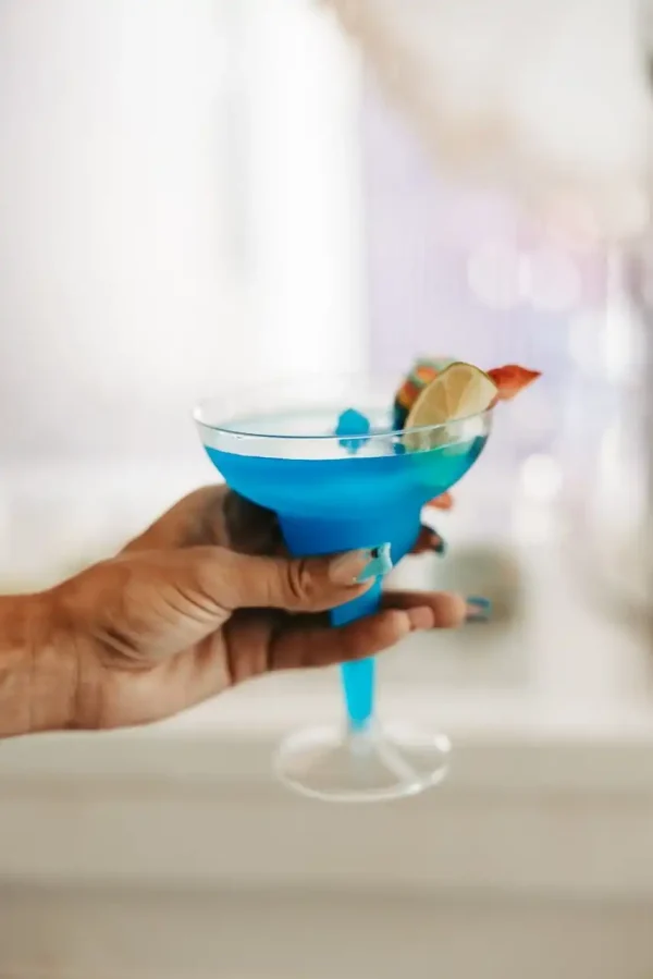 A hand holding a blue cocktail in a martini glass, garnished with a lime slice and a small orange decoration, perfect for showcasing in our Event Photography Packages.