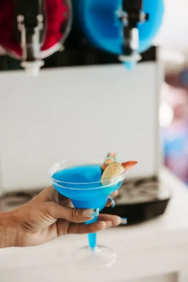 A hand holds a blue cocktail in a glass garnished with a lime wedge and a small paper umbrella at an event, making the scene perfect for capturing with our Event Photography Packages.