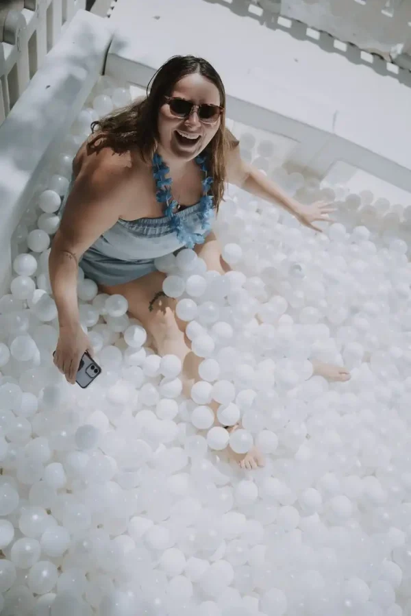 A woman wearing sunglasses and a blue lei sits in a pool filled with white plastic balls, smiling and holding a smartphone, perfectly capturing the joyful moment afforded by our Event Photography Packages.