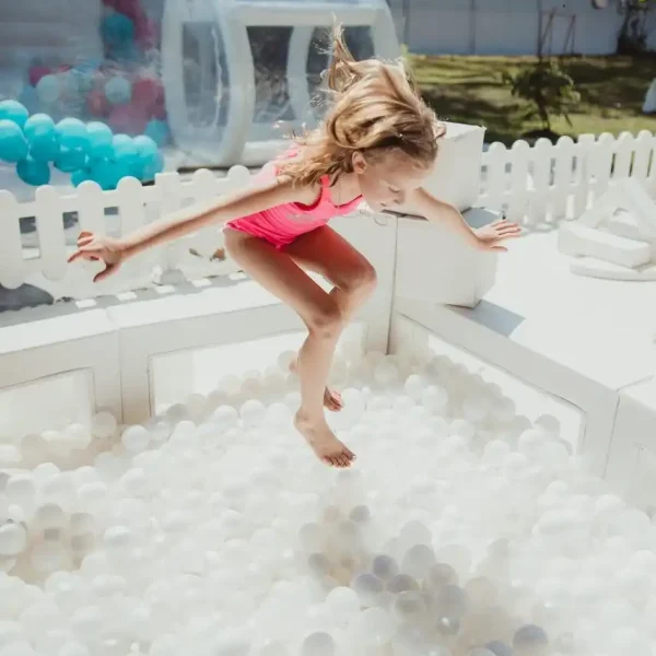 A child in a pink swimsuit leaps into a white ball pit in a soft play rental in outdoor play area, encircled by a fence and inflatable structures, embodying the joyful moments perfect for showcasing with our Event Photography Packages.