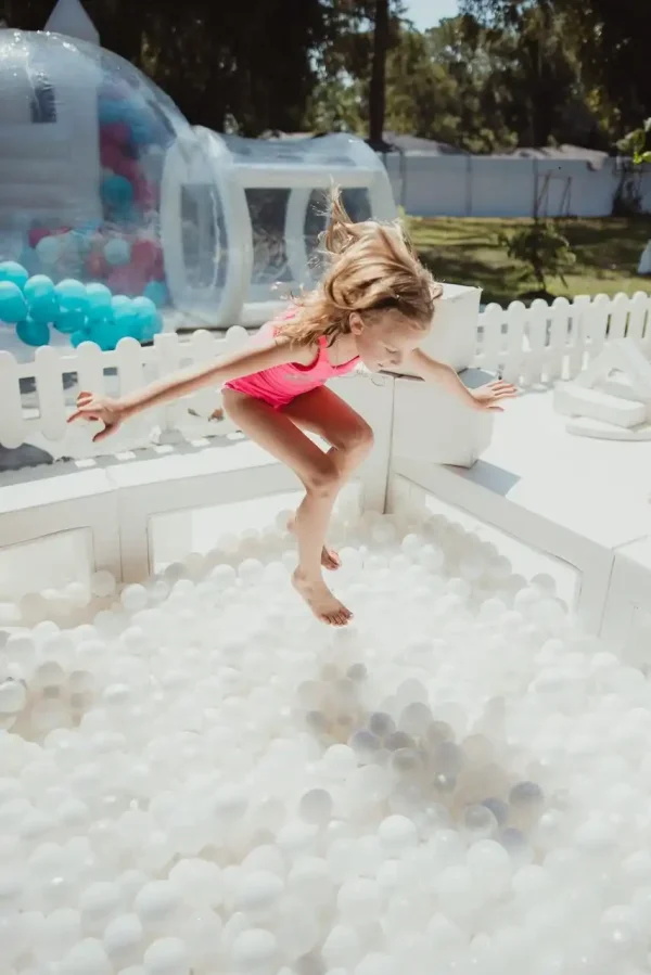 A child in a pink swimsuit leaps into a white ball pit in a soft play rental in outdoor play area, encircled by a fence and inflatable structures, embodying the joyful moments perfect for showcasing with our Event Photography Packages.