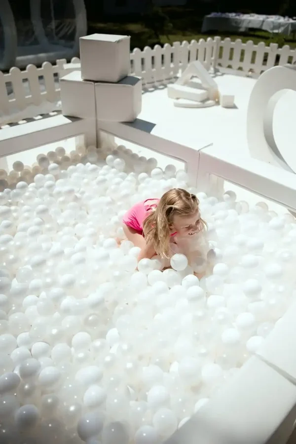 A child dressed in a pink outfit plays inside a white ball pit, enclosed by a white picket fence—perfectly exemplifying the joyful essence captured by our Event Photography Packages.