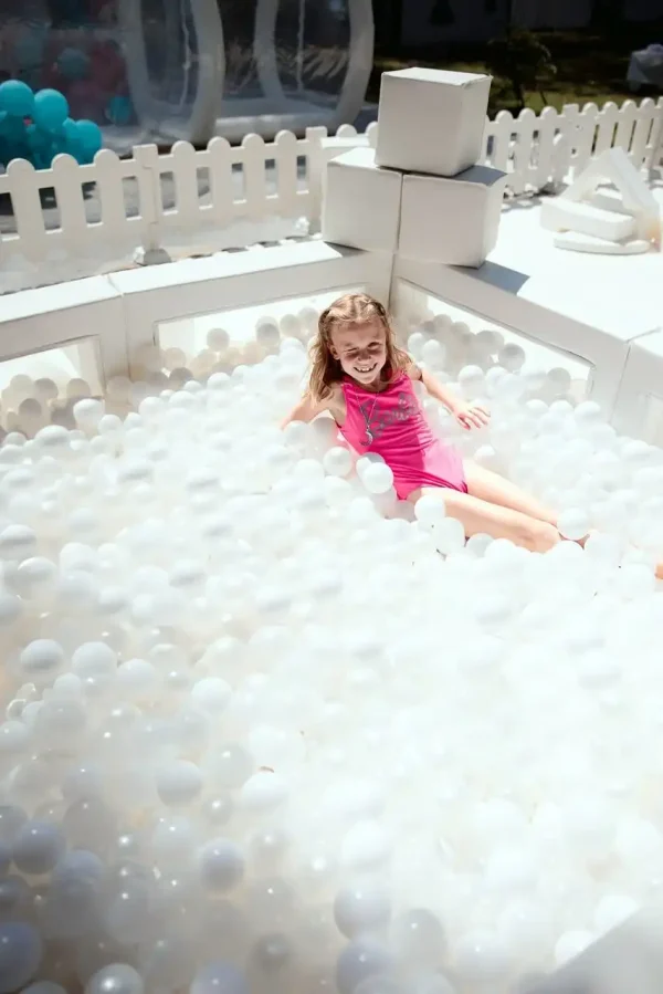 A young girl in a pink swimsuit sits in a ball pit filled with white plastic balls, surrounded by white fencing and blocks—perfect for capturing memorable moments with our Event Photography Packages.