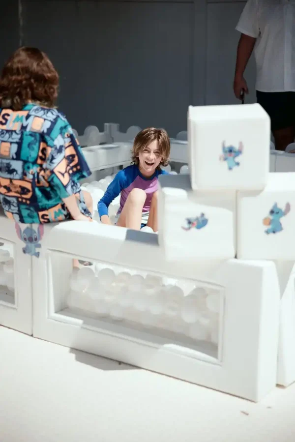 A child plays in a white play area with another child nearby, surrounded by white foam blocks featuring images of Stitch from "Lilo & Stitch." Capture these precious moments with our Event Photography Packages, ensuring every delightful memory is beautifully preserved.