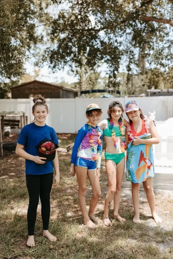 Four children stand together outside, three wearing colorful swimwear and one in a blue shirt and black pants holding a hat. With a fence and trees in the background, it’s the perfect moment to capture using our Event Photography Packages tailored for any occasion.