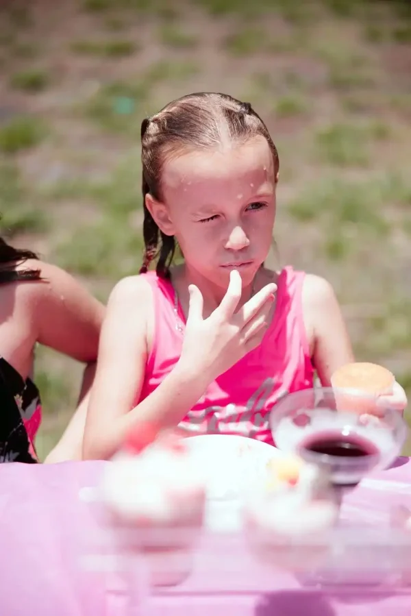 A young girl in a pink tank top licks frosting off her fingers while holding a cupcake, effortlessly captured by our Event Photography Packages at an outdoor event, sitting at a pink-covered table.