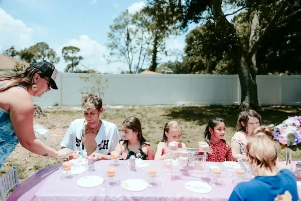 Several children sit at a table outdoors, set for a gathering with food and drinks, as an adult serves them. Trees and a white fence are in the background. This scene perfectly captures the essence of our Event Photography Packages, ideal for preserving such joyful memories.