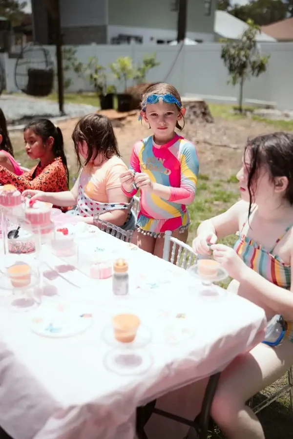 Children in swimsuits are seated around a table outdoors, immersed in arts and crafts activities on a sunny day—an ideal setting to capture unforgettable moments with our Event Photography Packages.