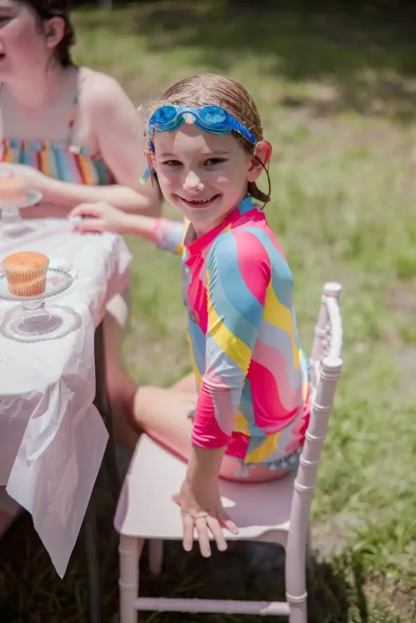 A smiling child in a colorful swimsuit and blue goggles sits on a white chair at an outdoor table enjoying cupcakes, perfectly showcasing our Event Photography Packages.