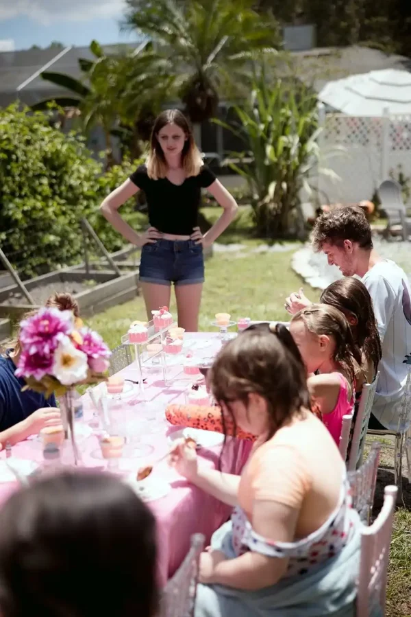 A woman stands with hands on hips beside a table where children are seated, eating, and engaged in activities at an outdoor gathering decorated with pink tablecloths and flowers, presenting the ideal scene for showcasing our Event Photography Packages.