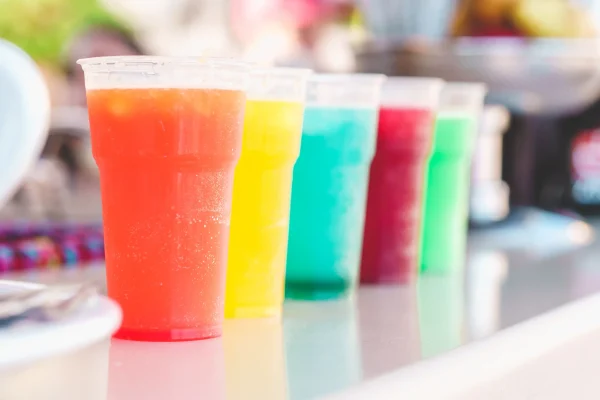 Five vibrant plastic cups, each filled with a different beverage, are arranged on a countertop in a manner that brings to mind the Slushy/Margarita Bar Package. The drinks are orange, yellow, blue, green, and red—any of which could effortlessly be a refreshing margarita or a lively slushy.