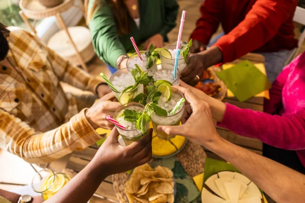 A group of people clink glasses with margaritas from the Slushy/Margarita Bar Package over a wooden table filled with tortilla chips and various dishes.