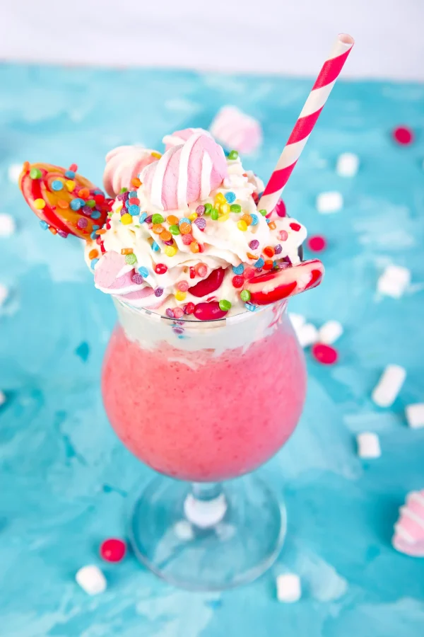 A tall glass of a pink slushy from the Slushy/Margarita Bar Package, topped with whipped cream, sprinkles, colorful candies, small meringues, and a red and white striped straw against a blue background—perfect for those craving a playful slushy experience.