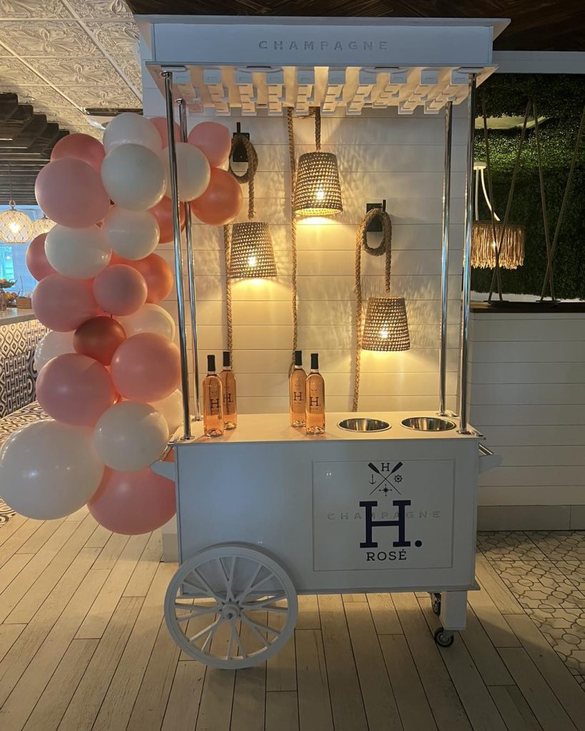 Discover the ultimate baby shower inspiration with this stunning white champagne cart, elegantly branded with an "H." emblem. The cart beautifully displays three exquisite bottles of rosé, perfectly positioned beneath an enchanting array of four wicker pendant lights that cast a warm glow over the scene. To the left, an eye-catching cluster of pink and white balloons elevates the festive ambiance, while a luxurious wooden floor and lush green plant wall backdrop add to the sophisticated charm of this premier Ultimate Baby Shower Rentals setup. Ideal for those seeking a memorable event adorned with chic decor and premium aesthetics.