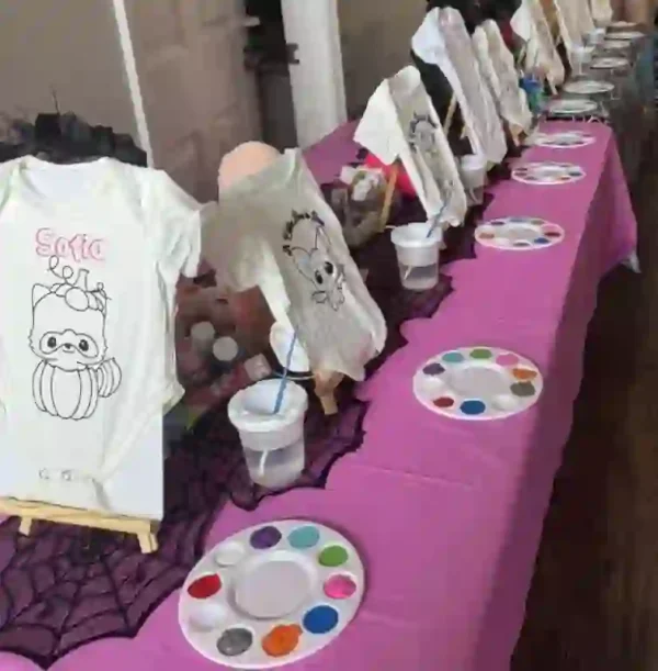 Experience an enchanting painting activity setup by Ultimate Baby Shower Rentals, perfect for a memorable baby shower event. The table, elegantly draped in a vibrant pink tablecloth, is adorned with a charming array of small easels displaying pristine white onesies decorated with delightful cartoon designs. Each seat is thoughtfully equipped with its own paint palette, complemented by jars of water and an assortment of brushes, inviting guests to unleash their creativity. This whimsical arrangement promises not only a fun and interactive experience but also serves as a picturesque backdrop for your baby shower celebration. Perfect for those seeking engaging baby shower ideas centered around artistic expression!