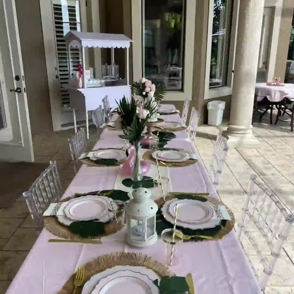 Discover the epitome of sophistication with this luxurious outdoor table setup, perfect for those searching for elegant event rentals. A soft pink tablecloth sets the stage, beautifully accented by shimmering gold cutlery and pristine white plates. Verdant green leaf decor adds a fresh touch, complementing the chic ambiance. Surrounding the table are sleek clear Adult Ghost Chairs, offering both comfort and style. In the background, a charming dessert cart adds a sweet finishing touch to this refined setting, ideal for upscale special occasions and glamorous gatherings.