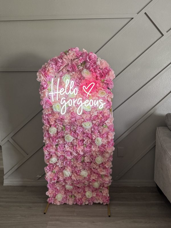 Capture the essence of elegance and charm with this eye-catching decorative sign featuring a stunning floral wall. Lush pink and white blossoms create a captivating backdrop, perfectly complementing the radiant neon sign that warmly greets with the words "Hello Gorgeous" alongside a dainty heart symbol. The flowers pop against the chic gray wall with contemporary diagonal lines, while part of a sleek light gray sofa enhances this stylish setting. Ideal for those seeking vibrant decor inspiration or Instagram-worthy photo backdrops, this arrangement combines modern aesthetics with floral beauty to create an inviting atmosphere.