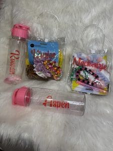 Engaging and SEO-optimized alt text: Vibrant clear plastic water bottles labeled "Rowan" and "Aspen," each topped with playful pink lids, rest on a luxurious white fluffy surface, creating an inviting scene. Nearby, two transparent bags adorned with the name "Mackenzie" showcase an assortment of vibrant plastic beads, a whimsical multicolored pompom garland, and enchanting flower accessories. A versatile bottle of acrylic sealer stands ready to add the finishing touch to your creative projects. Perfect for DIY enthusiasts or personalized gifts ideas!