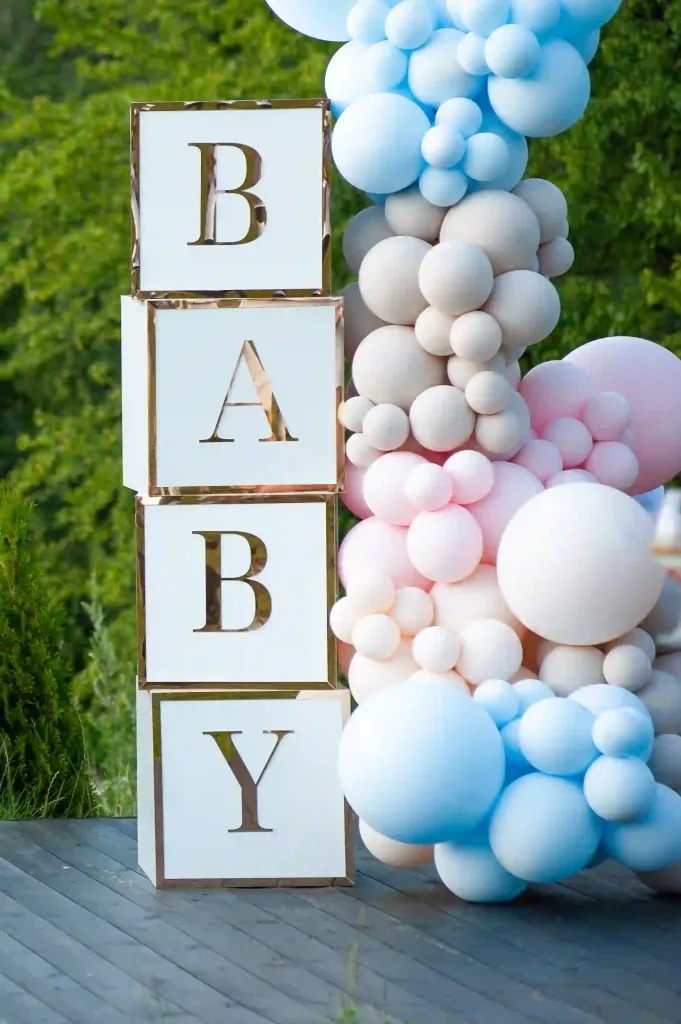 Experience the perfect outdoor baby shower setting with a stunning vertical display of four large blocks spelling "BABY" in bold black letters against a crisp white background. These chic blocks are elegantly surrounded by vibrant clusters of balloons in delightful shades of pink, blue, and beige. The lush green foliage provides a picturesque backdrop, enhancing the welcoming and festive atmosphere for your ultimate baby shower rentals event. Perfect for capturing unforgettable memories at your dream celebration.