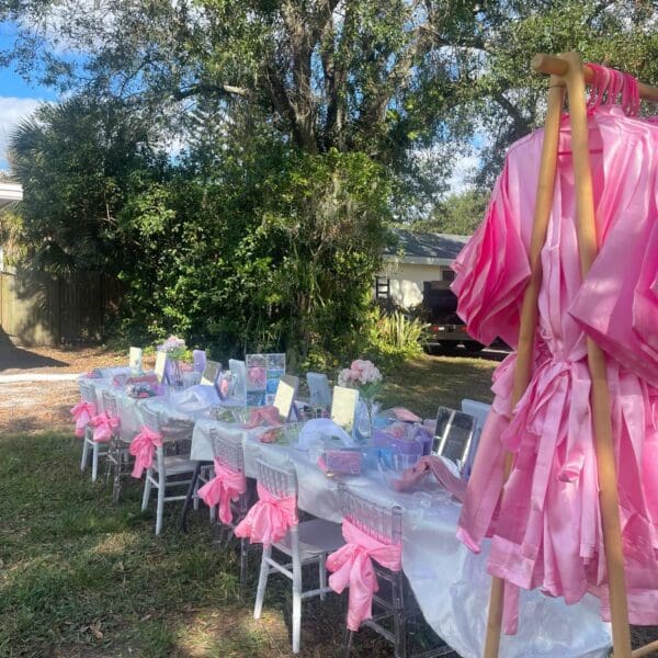 Captivating outdoor event setup with enchanting pink-themed decor. An elegant long table is beautifully adorned with delicate pink bows and meticulously arranged place settings, creating a whimsical ambiance. Nearby, a wooden rack elegantly displays luxurious pink robes, offering a touch of sophistication. Lush trees and vibrant foliage provide a serene and picturesque backdrop, enhancing the charm of this delightful gathering space. Perfect for garden parties and chic outdoor celebrations!