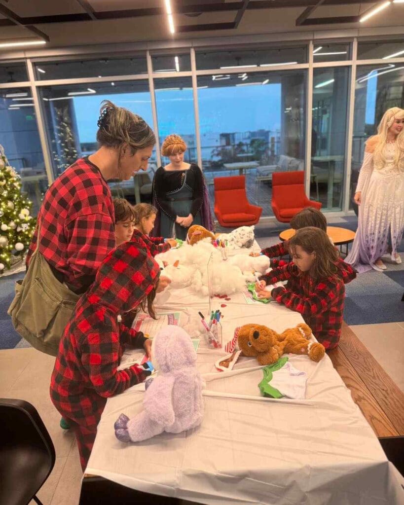 Delightful scene of children in festive red plaid pajamas joyfully decorating plush toys at a table, closely guided by adults dressed in charming holiday costumes. A beautifully adorned Christmas tree glittering with ornaments and twinkling lights stands majestically in the background, adding a touch of seasonal magic to this heartwarming family activity. Perfect keywords: festive family activity, holiday crafting session, children decorating toys, cozy Christmas gathering.