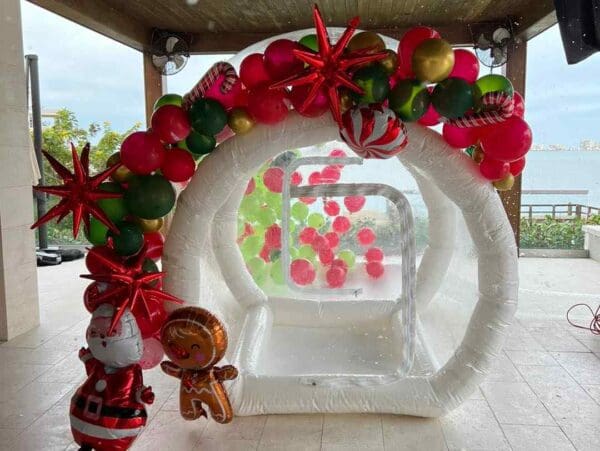 Discover the ultimate festive atmosphere with this enchanting inflatable white igloo, ideal for your holiday parties. Adorned with vibrant red, pink, and green balloons alongside dazzling star-shaped ornaments, this eye-catching centerpiece promises to captivate guests. Flanking the entrance are two cheerful character inflatables that add an extra touch of whimsy and delight. All of this is set against a breathtaking ocean view backdrop, offering a picturesque setting for unforgettable celebrations. Perfect for creating magical moments full of holiday cheer!