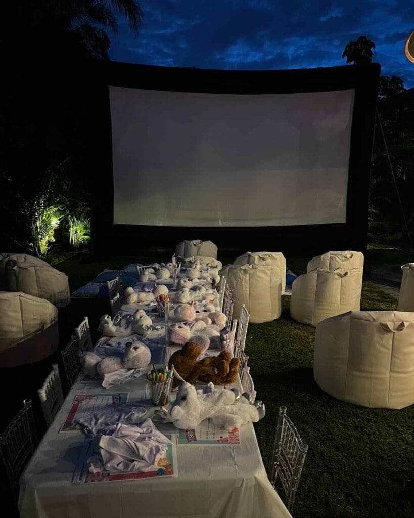 Experience the ultimate outdoor movie night with a captivating setup featuring a massive screen under the starlit sky. Plop down on comfy bean bags arranged for perfect viewing, surrounded by nature's beauty with tall trees swaying in the gentle breeze. A charming table nearby brims with adorable stuffed animals and vibrant coloring materials, adding a playful touch for family-friendly fun. Perfect your evening in this enchanting setting that combines entertainment and creativity in one delightful package. Perfect keywords: outdoor movie setup, large screen, family-friendly entertainment, scenic natural backdrop, creative activities.