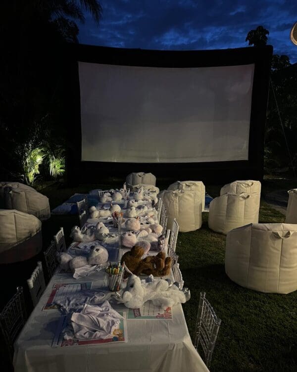 Immerse yourself in a magical outdoor movie night setup featuring a massive, high-definition screen set against the backdrop of a stunning evening sky. Experience ultimate comfort with plush white bean bags strategically arranged for the perfect view. A charming table centerpiece boasts an array of adorable stuffed animals and vibrant art supplies, perfect for creative moments before the film begins. Enjoy this enchanting cinematic experience under the stars, ideal for family gatherings or memorable date nights in your backyard oasis.