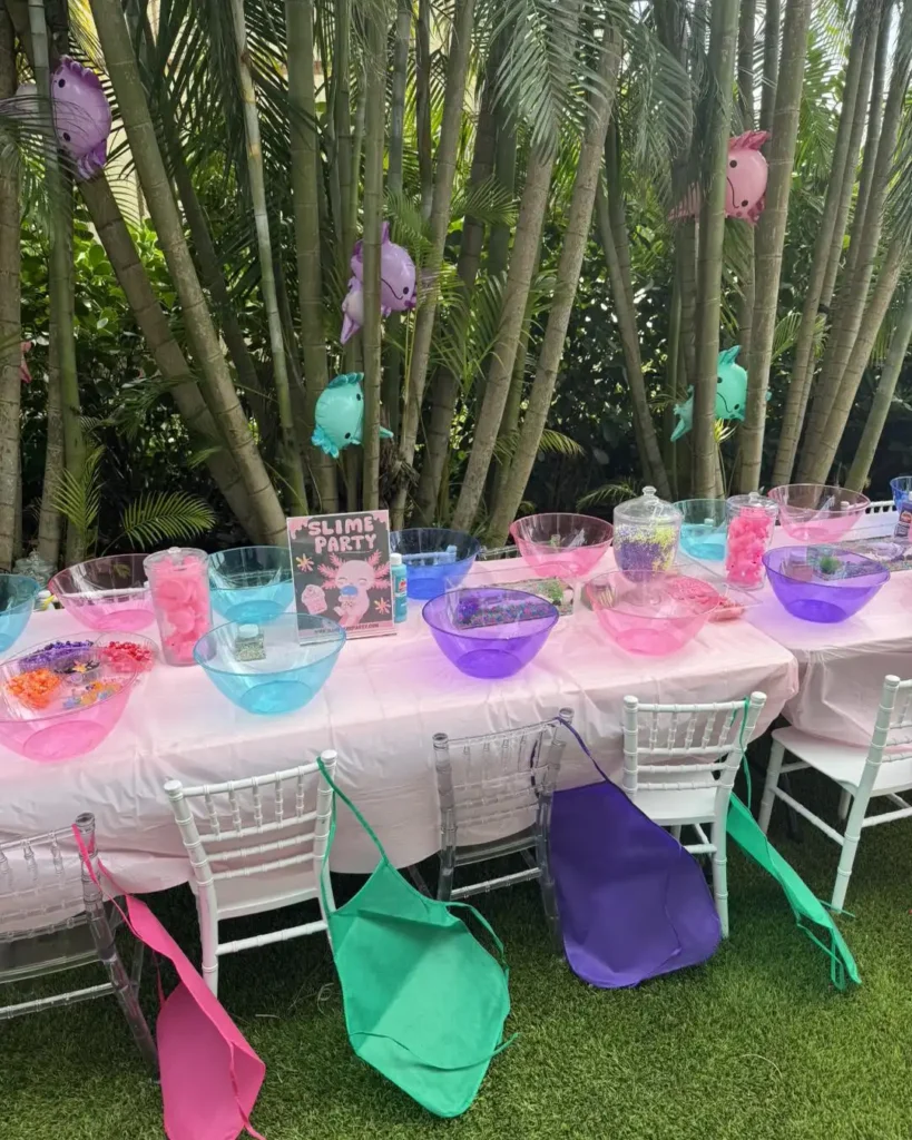 Engaging and colorful outdoor mobile slime-making party setup, inviting creativity and fun for corporate gatherings. A long table is elegantly arranged with vibrant bowls brimming with an array of colorful ingredients, ready for guests to craft their own slime masterpieces. Crisp white aprons drape over chic white chairs, enticing attendees to dive into this hands-on experience. The backdrop features swaying palm trees and whimsical decorative balloons, adding a festive and tropical flair that elevates this unique corporate party idea into an unforgettable event.