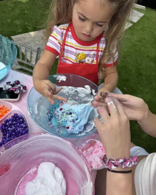 A young child in a vibrant red apron enthusiastically stirs a kaleidoscope of colorful beads into shimmering blue slime at an outdoor table, capturing the joy and creativity of a lively slime-making party under the open sky. Perfect for sensory play enthusiasts, this dynamic scene highlights the imaginative fun and hands-on excitement that make slime parties a memorable experience for kids.