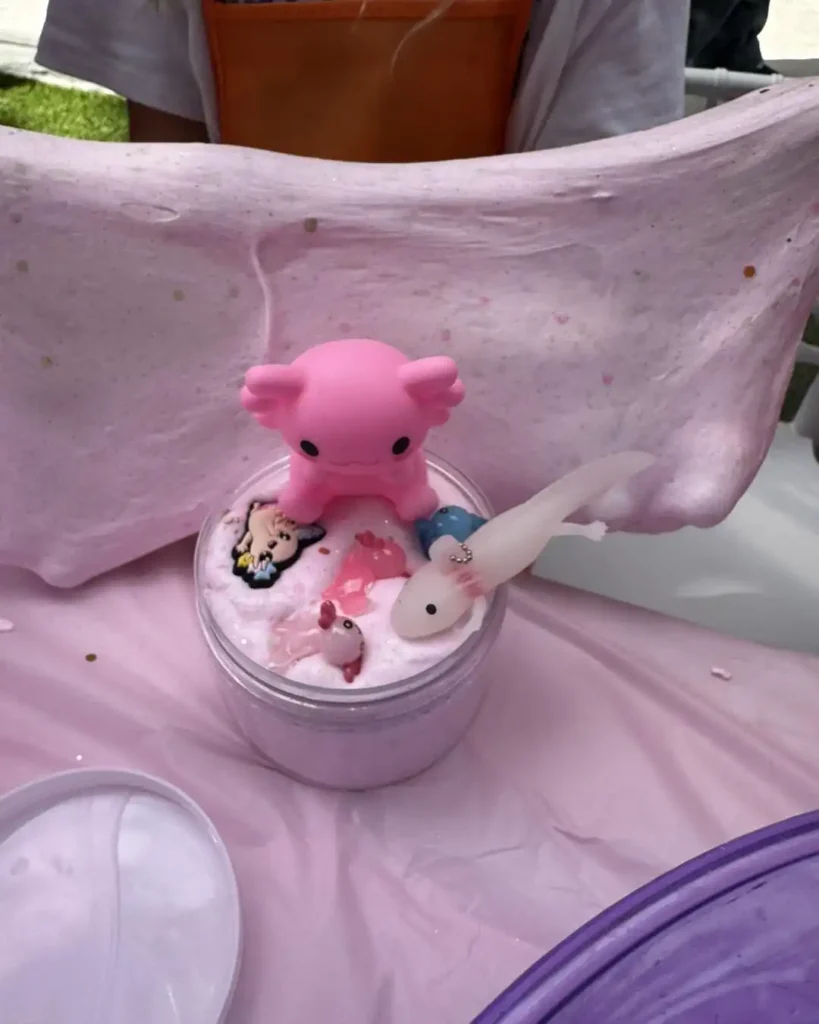 Lively Mini Slime Party with Cute Pink and White Axolotl Toys: Discover a whimsical world of slime fun as a vibrant pink container of glossy slime becomes the center stage for an enchanting mini party! A charming pink axolotl toy sits playfully atop the gooey surface, while its adorable white companion waits eagerly beside it. In the background, hands expertly stretch and swirl the stretchy slime, creating an engaging sensory experience that's both visually captivating and fun. Perfect for children's entertainment or satisfying ASMR enthusiasts, this scene captures the magic of creative playtime through colorful textures and delightful characters.