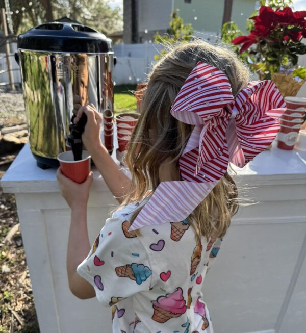 A young girl, adorned in a whimsical shirt featuring vibrant ice cream cones and charming heart patterns, eagerly pours a refreshing drink from a large dispenser into a striking red cup. Her outfit is completed with an oversized bow, combining playful pink and crisp white hues, adding an extra layer of cuteness to the scene. This moment evokes delightful memories of summer adventures at the nearby refreshment stand beside the bustling Snow Machine Rental area. Perfect for those seeking unique hydration options amidst snowy fun!