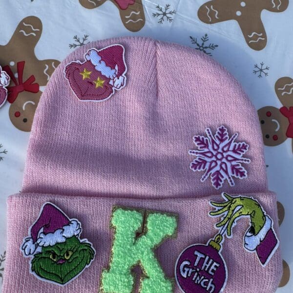 Vibrant pink beanie embellished with festive Christmas-themed patches, perfect for winter enthusiasts. This cozy hat showcases a delicate snowflake design, the initial "K," and an adorable smiling green face donned in a Santa hat. A whimsical hand holding a red ornament marked "The Grinch" completes the charming detail. Photographed against an enchanting backdrop of gingerbread patterns, this beanie is your must-have winter accessory—ideal for keeping warm on snowy days after exhilarating adventures with a snow machine rental. Let seasonal cheer elevate your style this holiday season!