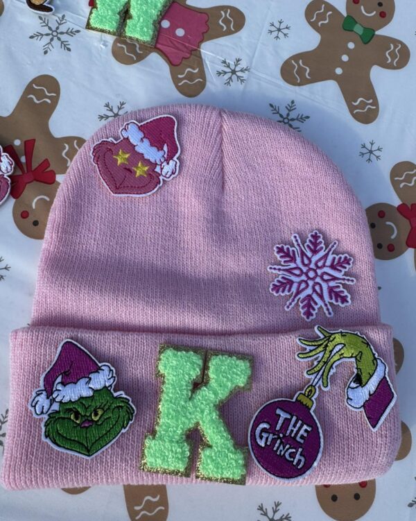 Vibrant pink beanie embellished with festive Christmas-themed patches, perfect for winter enthusiasts. This cozy hat showcases a delicate snowflake design, the initial "K," and an adorable smiling green face donned in a Santa hat. A whimsical hand holding a red ornament marked "The Grinch" completes the charming detail. Photographed against an enchanting backdrop of gingerbread patterns, this beanie is your must-have winter accessory—ideal for keeping warm on snowy days after exhilarating adventures with a snow machine rental. Let seasonal cheer elevate your style this holiday season!