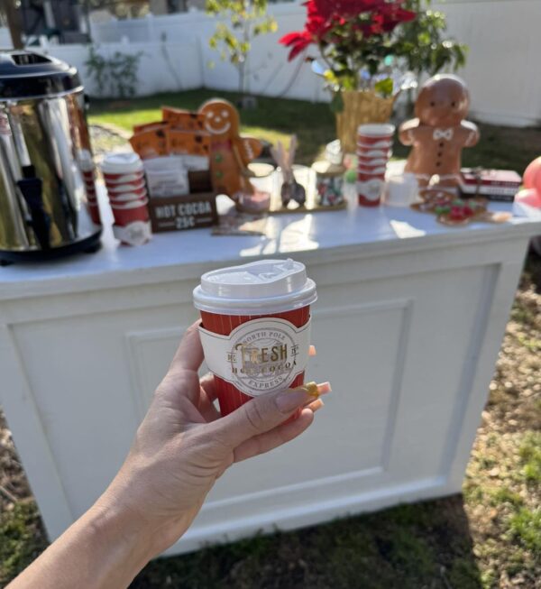 A cozy hand holds a steaming cup of coffee amidst a charming outdoor holiday setup, featuring a rustic wooden stand decked out with hot cocoa delights, festive gingerbread cookies, and cheerful seasonal decorations. In the background, the nearby Snow Machine Rental adds an authentic wintry touch to this enchanting Christmas market scene. Perfect for winter wanderlust enthusiasts seeking heartwarming moments and picturesque backdrops!