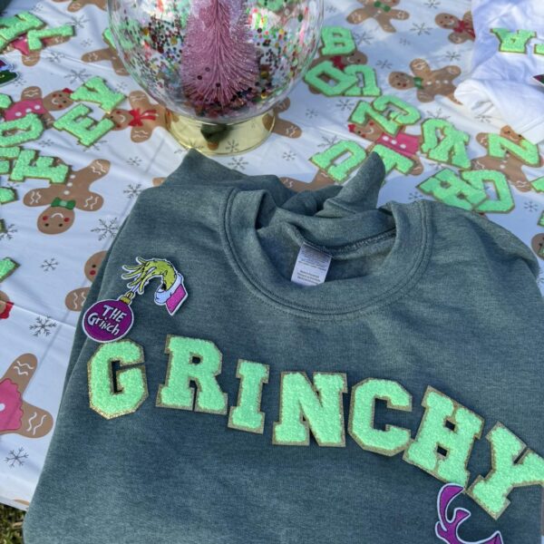 A vibrant green sweatshirt emblazoned with bold "GRINCHY" lettering lies prominently on a table, perfectly capturing the playful essence of holiday cheer. Nestled beside this cozy attire is a dazzling pink glittery tree ornament, exuding festive sparkle and charm. Complementing the scene is whimsical gingerbread-themed decor, adding a touch of warmth and sweetness to your holiday aesthetic. Elevate your seasonal celebration with this enchanting setup that's ideal for any holiday party. Enhance the wintry ambiance even further with the magical touch of a Snow Machine Rental, creating an unforgettable festive experience perfect for sharing on social media!