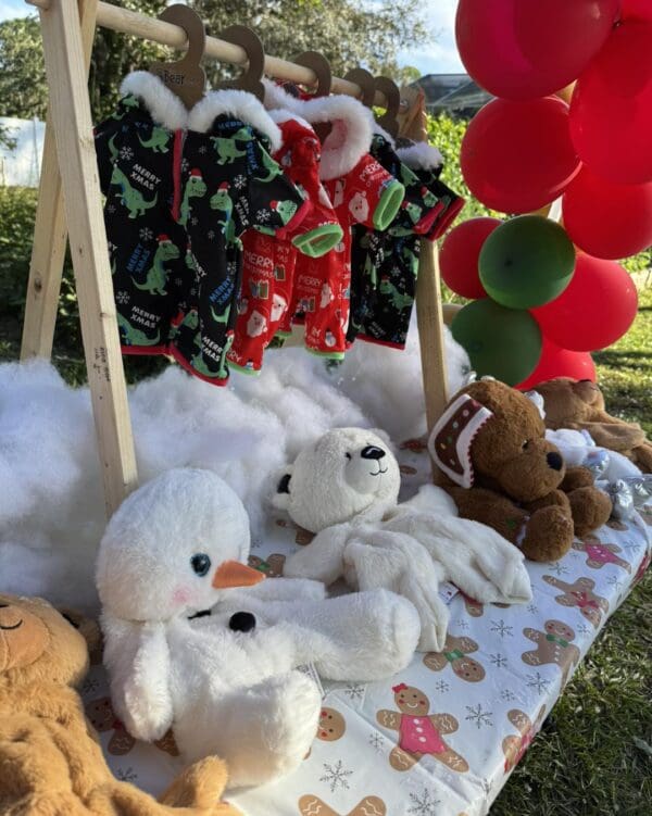A whimsical display of festive-themed children's clothing hangs invitingly on charming wooden hangers, featuring vibrant colors and playful patterns perfect for holiday celebrations. Below the eye-catching apparel, an enchanting arrangement of cuddly plush toys sits nestled amidst a fluffy layer of cotton snow, delicately crafted by the renowned Snow Machine Rental. Surrounding this magical scene are bursts of cheerful, colorful balloons that add extra sparkle to the joyful atmosphere. This captivating setup perfectly captures the essence of a winter wonderland while highlighting seasonal fashion trends for children and creating an engaging shopping experience. Ideal for anyone seeking unique holiday gift ideas or inspiration in festive kids' fashion displays!