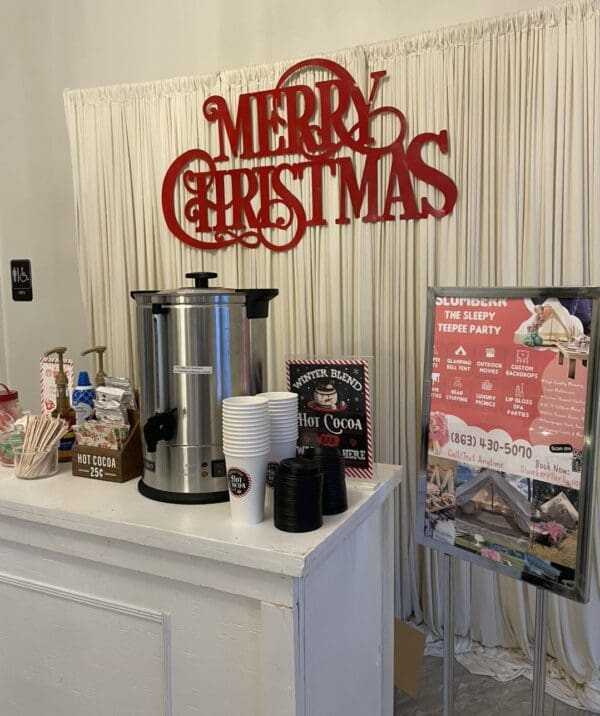 Step into a winter wonderland with the Movie Concession Stand Rental magically transformed into an inviting hot cocoa haven. This festive setup, elegantly situated on a pristine white counter, features a state-of-the-art cocoa dispenser, an array of cups, and delightful toppings that cater to every sweet tooth. Above the station hangs a charming "Merry Christmas" sign in bright holiday colors, instantly evoking the spirit of the season. To the right, an informative sign adds to the cheerful decor while offering insights into this seasonal treat experience. Perfect for holiday gatherings and events seeking that warm and cozy vibe!.