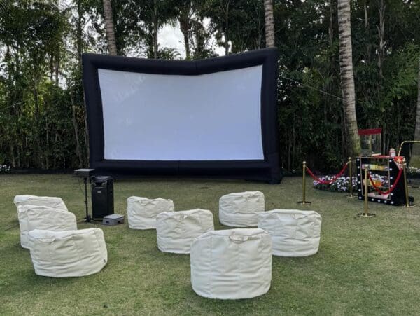 Experience the ultimate outdoor movie night with a captivating inflatable movie screen set against a lush backdrop of towering trees and soft grass. Enjoy a cozy ambiance as you sink into comfortable bean bag seating, perfectly arranged for optimal viewing pleasure. A charming small table is stocked with delicious popcorn and a high-quality speaker, enhancing your cinematic experience. Nearby, the convenient Movie Concession Stand Rental takes care of all your snacking desires effortlessly, providing an array of tasty treats and refreshments. Perfect for family gatherings or events under the stars, this setup transforms any outdoor space into an enchanting open-air theater experience.
