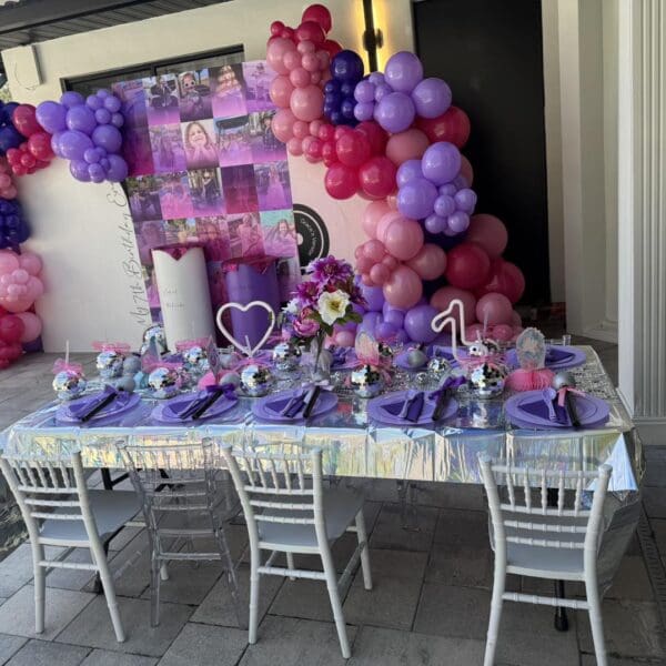 Experience the ultimate outdoor party setup in stunning cinematic style! A long table elegantly draped with a shimmering silver tablecloth serves as the centerpiece, complemented by rich purple napkins for a touch of sophistication. Lush floral centerpieces reminiscent of movie concession stand rentals add an artistic flair. The scene is brought to life by a vibrant balloon arch featuring lively shades of pink, purple, and red, creating the perfect backdrop for a memorable event under the sky. Perfect keywords: outdoor party setup, cinematic style, elegant table setting, floral centerpieces, vibrant balloon arch.