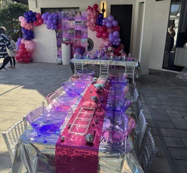 Discover the ultimate cinematic experience with a Movie Concession Stand Rental, beautifully transformed into a visual feast with vibrant pink, purple, and silver decorations. The inviting table display features elegant clear bowls set against a striking backdrop of eye-catching pink and purple balloons. This festive arrangement exudes irresistible charm, making it an ideal centerpiece for any party or movie-themed event. Perfect for drawing attention at your next celebration with its captivating color scheme and enchanting cinematic appeal!.