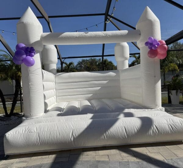 Experience the enchantment of a whimsical white inflatable bounce house adorned with vibrant clusters of purple and pink balloons, creating a captivating focal point on a sunlit patio. The clear enclosure overhead allows natural light to illuminate the festive setup, while palm trees sway gracefully in the background, evoking a tropical paradise. This picturesque setting provides the perfect backdrop for an unforgettable Movie Concession Stand Rental experience, where fun and relaxation blend seamlessly under blue skies. Ideal for outdoor events or parties, this Instagram-worthy scene invites guests to indulge in every moment of joy and entertainment amidst nature's beauty.