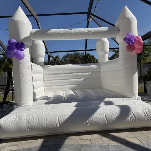 Experience the enchantment of a whimsical white inflatable bounce house adorned with vibrant clusters of purple and pink balloons, creating a captivating focal point on a sunlit patio. The clear enclosure overhead allows natural light to illuminate the festive setup, while palm trees sway gracefully in the background, evoking a tropical paradise. This picturesque setting provides the perfect backdrop for an unforgettable Movie Concession Stand Rental experience, where fun and relaxation blend seamlessly under blue skies. Ideal for outdoor events or parties, this Instagram-worthy scene invites guests to indulge in every moment of joy and entertainment amidst nature's beauty.